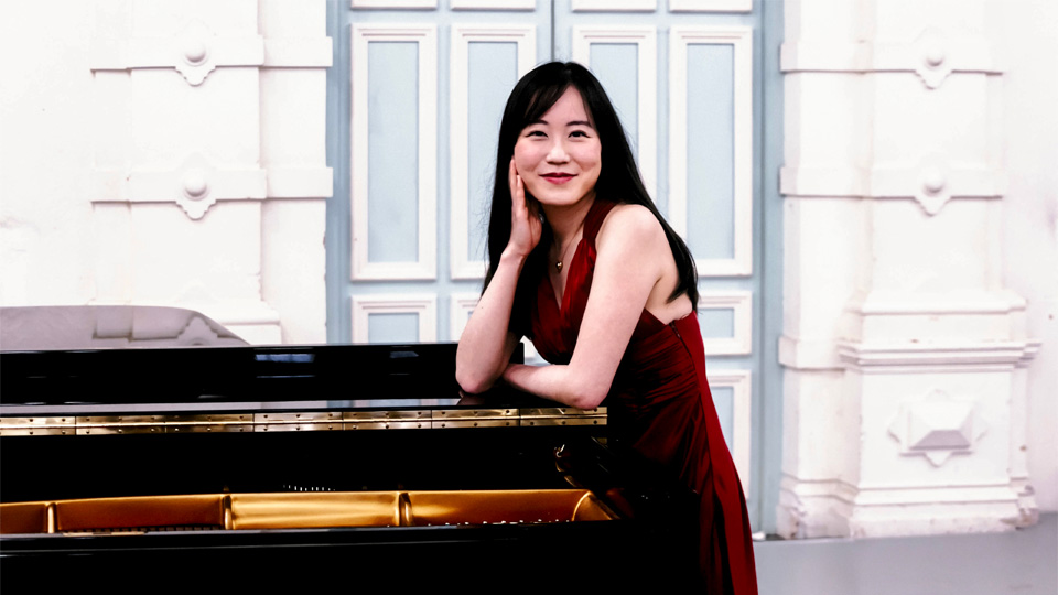 An Asian female student, with long dark hair, wearing a red dress, leaning against a piano, smiling at the camera.